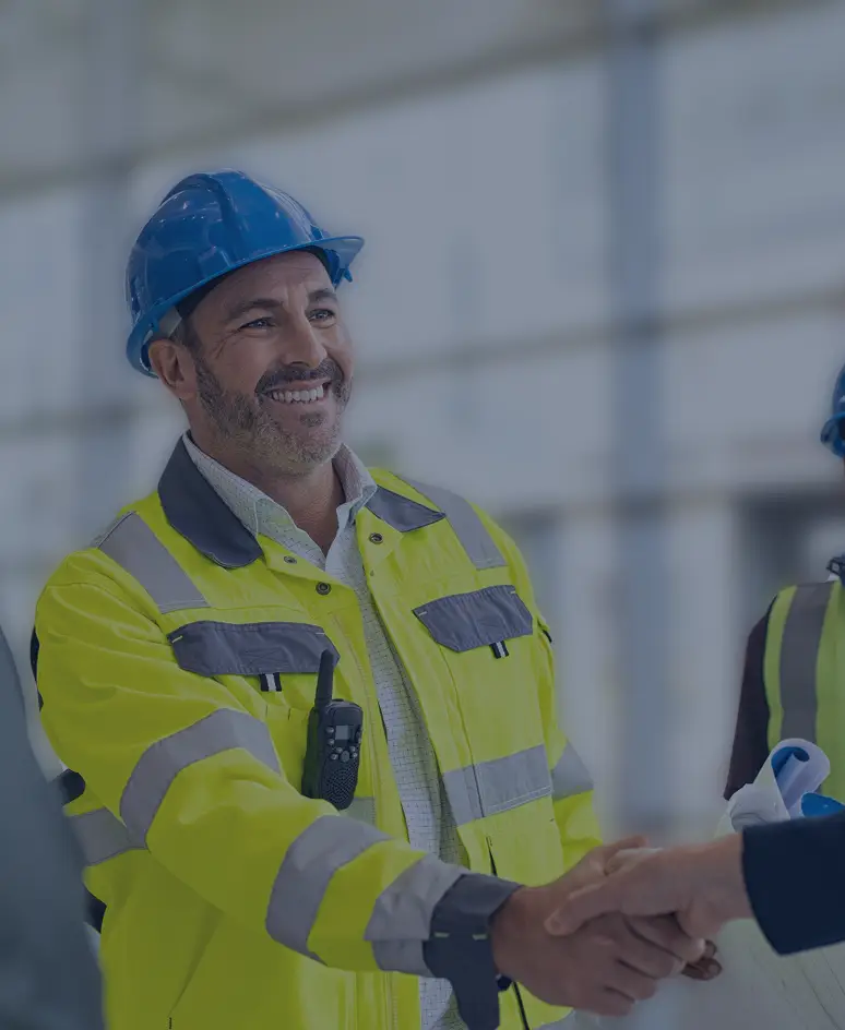 Safety officer in a high-visibility jacket and blue helmet smiling and shaking hands with a colleague inside a modern building, while other team members wearing safety gear look on.