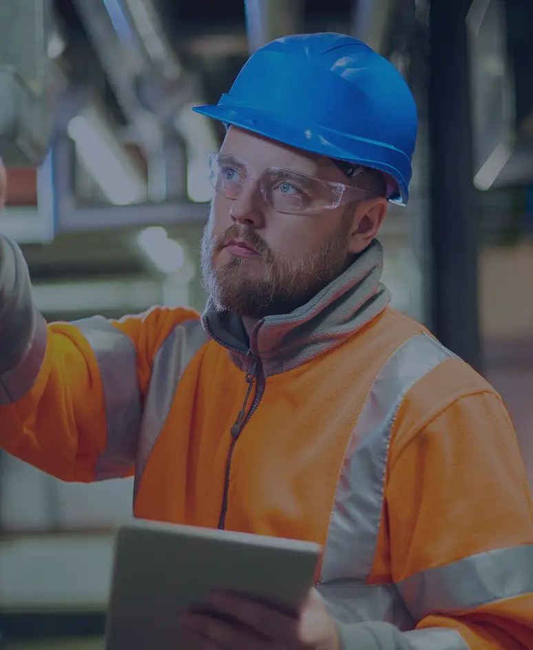 Safety officer wearing a blue helmet, safety glasses, and a high-visibility orange jacket inspecting a control panel while holding a digital tablet inside an industrial facility.
