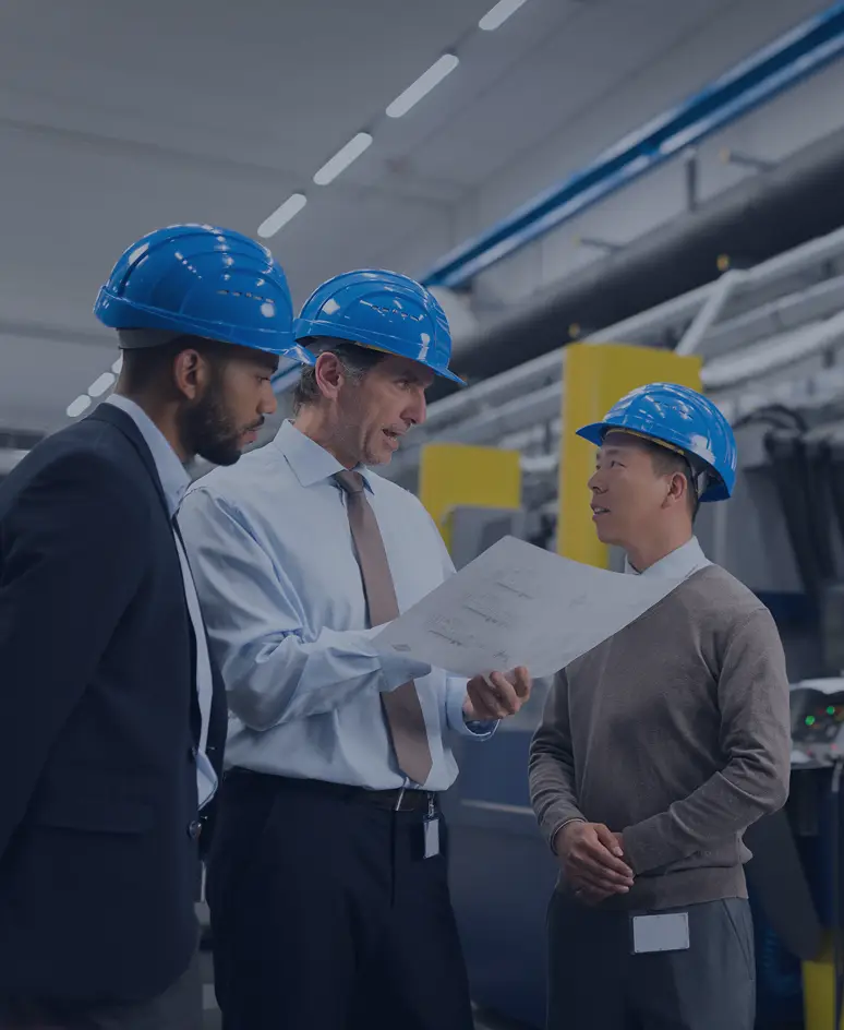Three safety officers wearing blue helmets reviewing blueprints inside a modern manufacturing facility, with industrial machinery in the background.